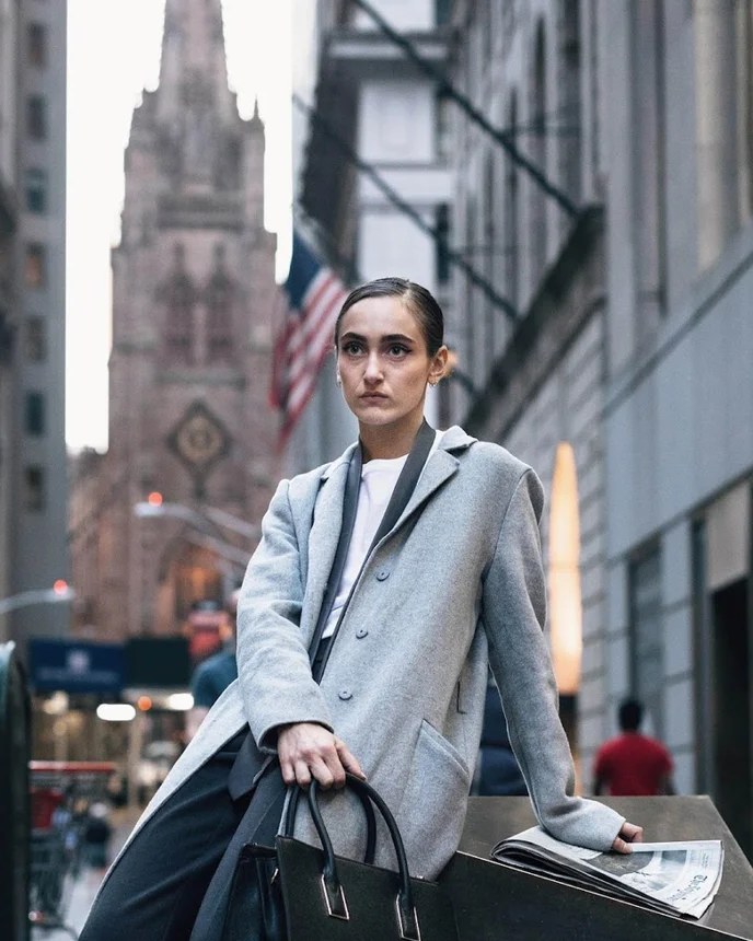 The SH-E-O: model in a grey coat on Wall Street, Trinity Church and an American flag behind her